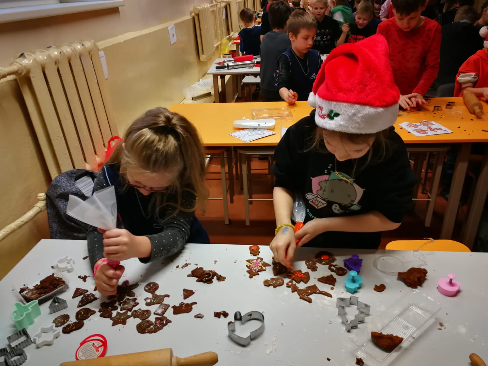 Divas meitenes sēž pie galda un veido piparkūkas ar formiņām. uz galda jau gatavas piparkūkas un formiņas.