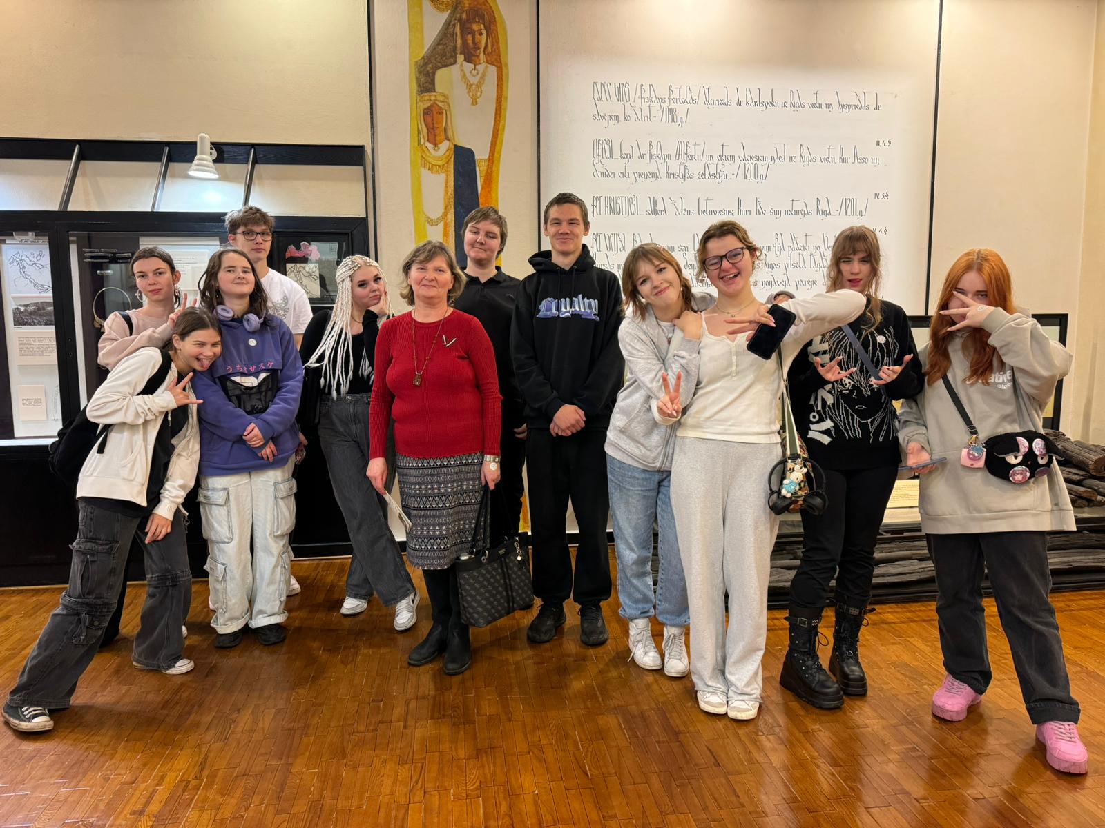 Visa klase, ar skolotāju sarkanajā džemperī muzeja telpās sastājusies un pozē fotogrāfam. Jaunieši smaida. Aiz muguras attēls ar dāmām un veco pierakstu.