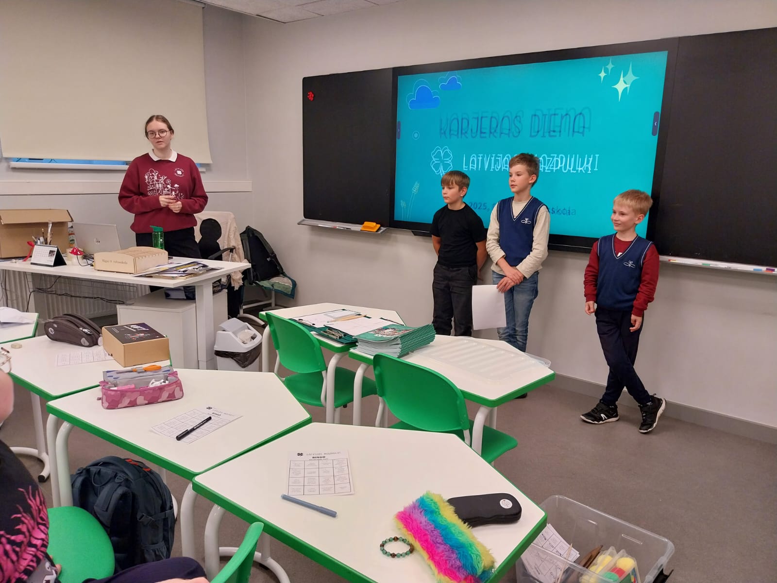 trīs skoleni stāv mpie tāfeles, kur mazpulku informacija, vadītaja jautā skolēniem.