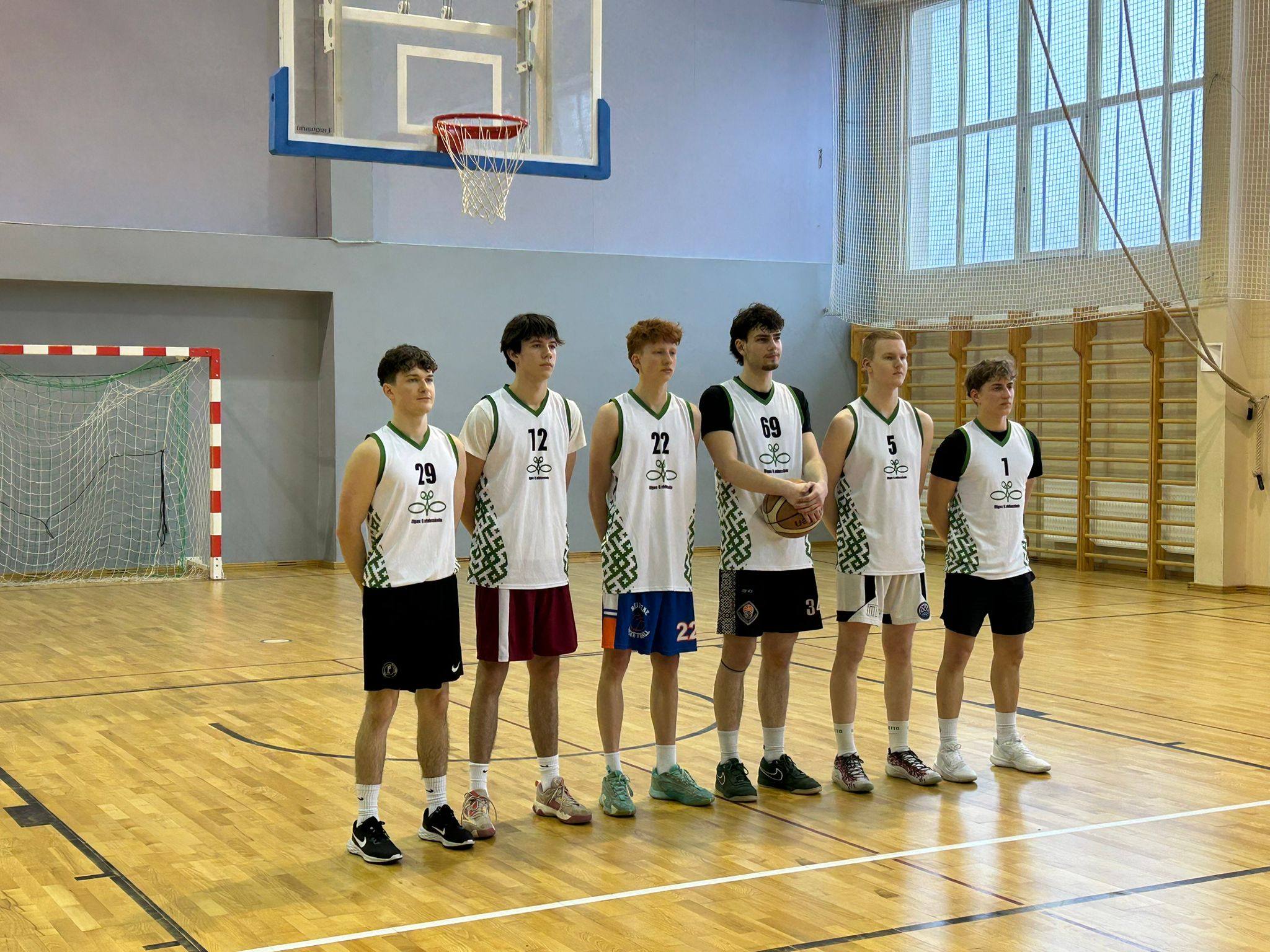 6 komandas puiši baltajos kreklos un melnajās šortos stāv sporta zālē