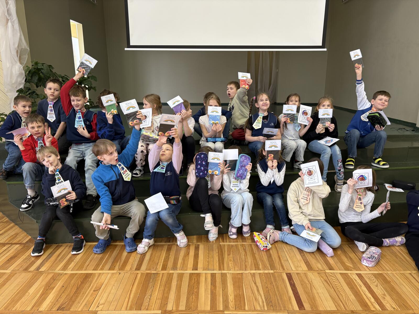 1. klašu skolēni vairākās rindās sēz uz skatuves, priecīgi, rokās tur diplomiņus