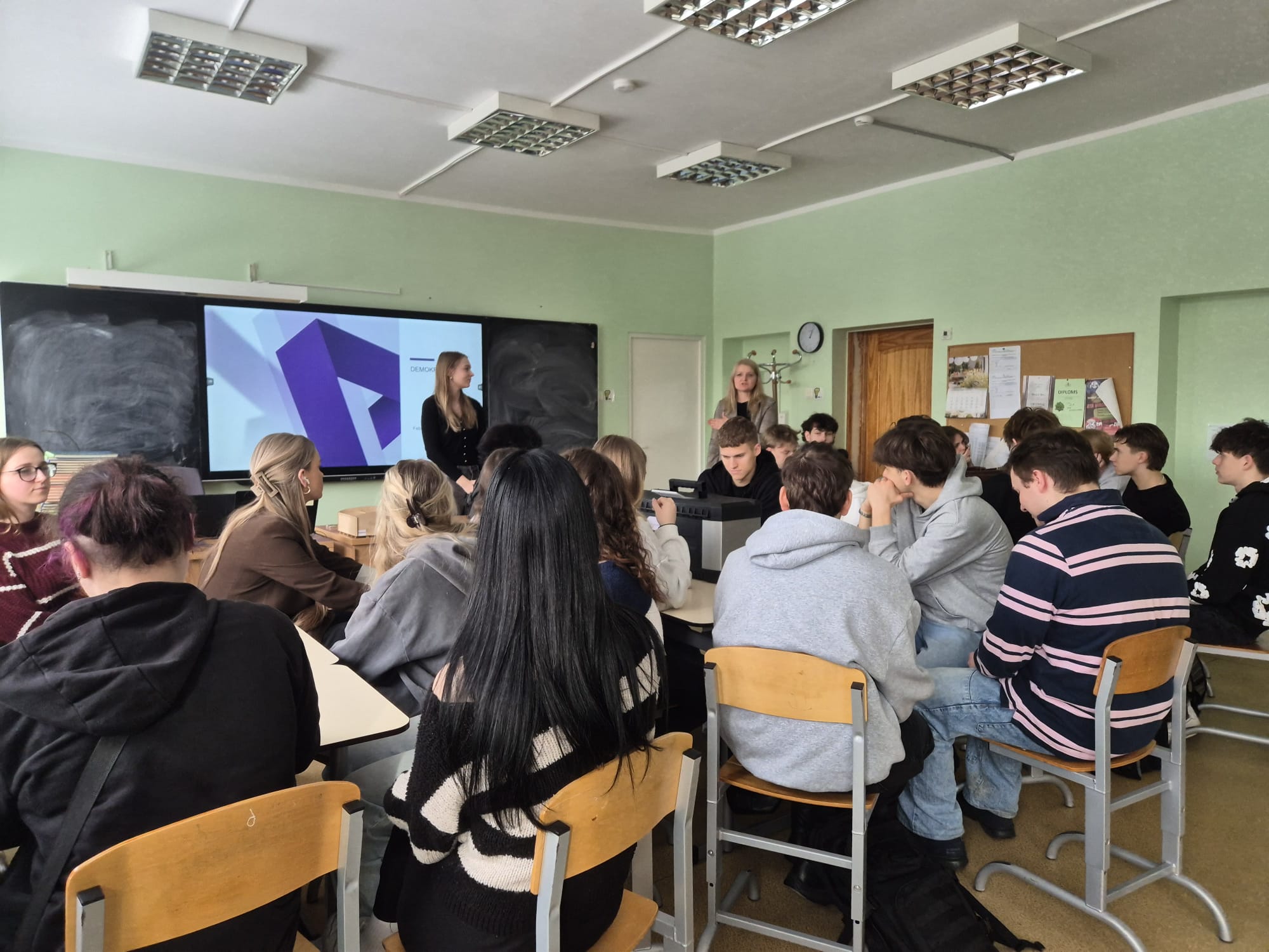 Daudz skolenu sēz pie galdiem, priekšā tāfele ar informāciju, lektore stāsta