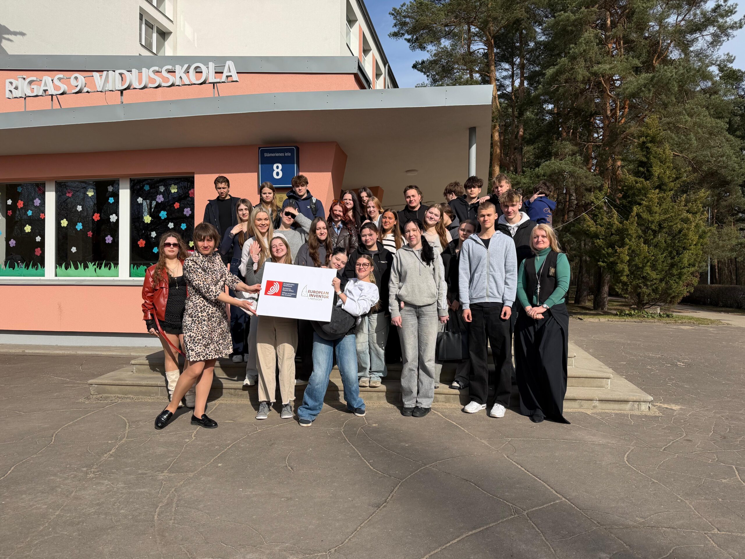 Jaunieši stāv skolās priekšā uz trepēm vairākās rindās, semināra vadītāja turplakātu.