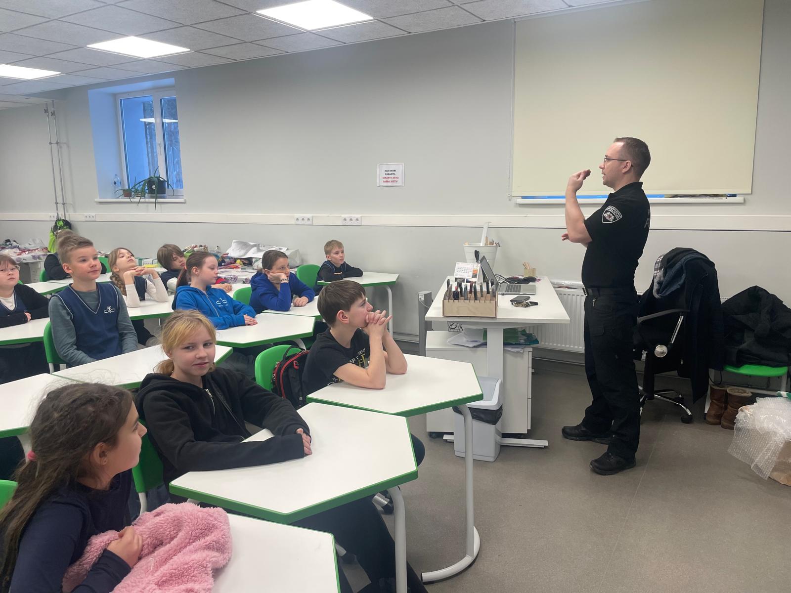 vīrietis policista tērpā stāsta jauniešiem, kas sēž skolas solos.