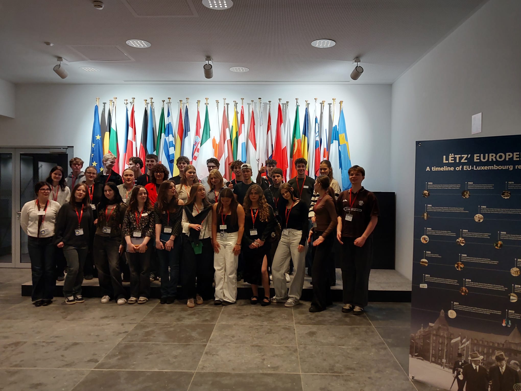 Skoleni stāv vairākās rindās, aiz muguras dažādu valstu karogi.