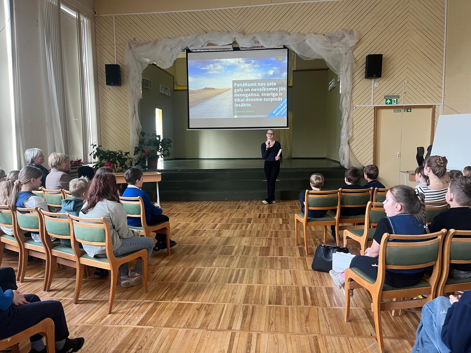 Titulbilde-lasīšanas konkurss- pie ekrāna zālē stāv meitene ar mikrofonu, skolēni sēž krēslos zālē