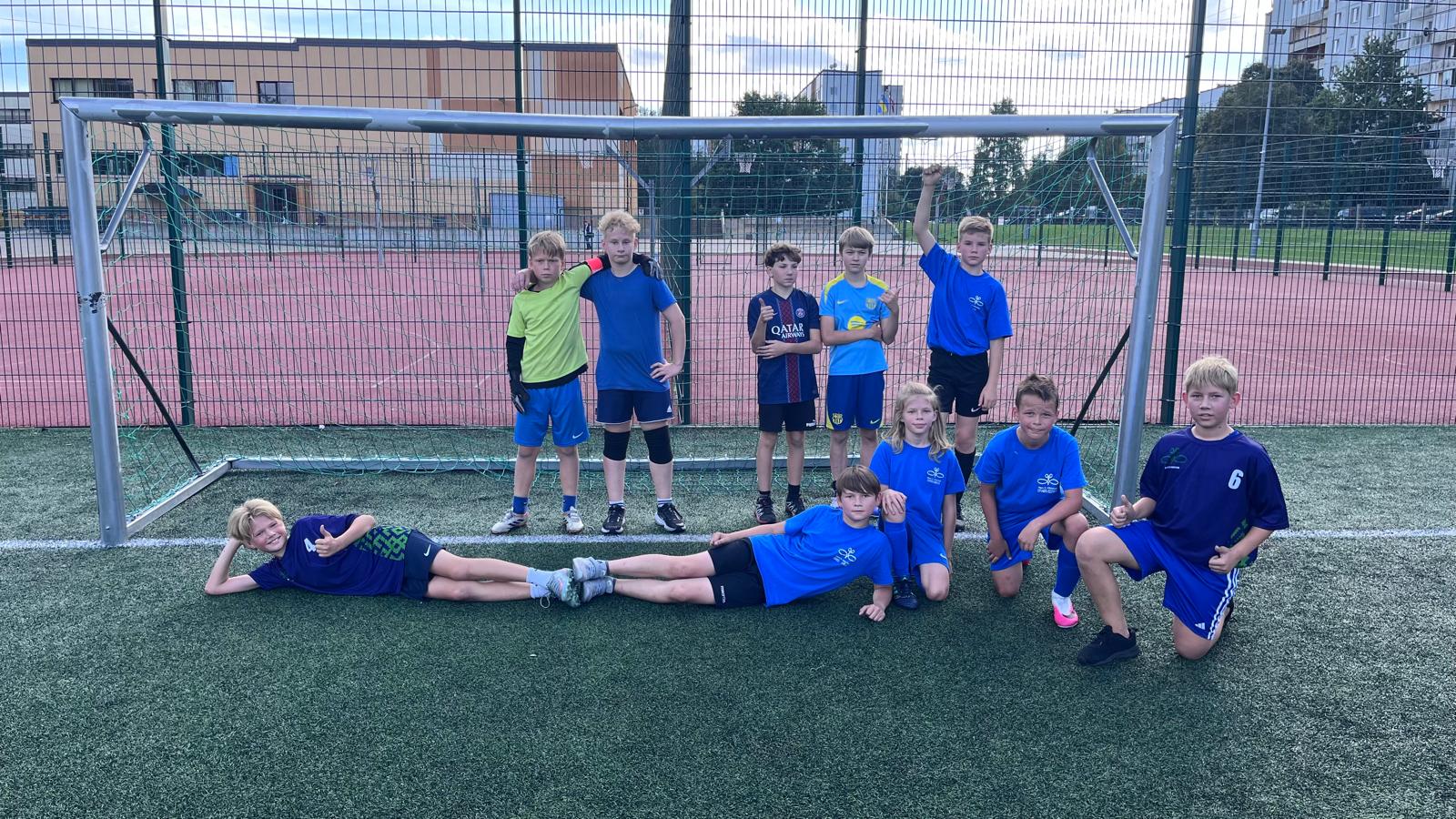 Mūsu skolas komandas puiši zilos krekliņos i stāv m, daži pietupusies pie futbola vārtiem.