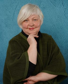 sieviete gaišiem matiem un tumso apmet, pielikusi roku pie zoda, nedaudz smaida.